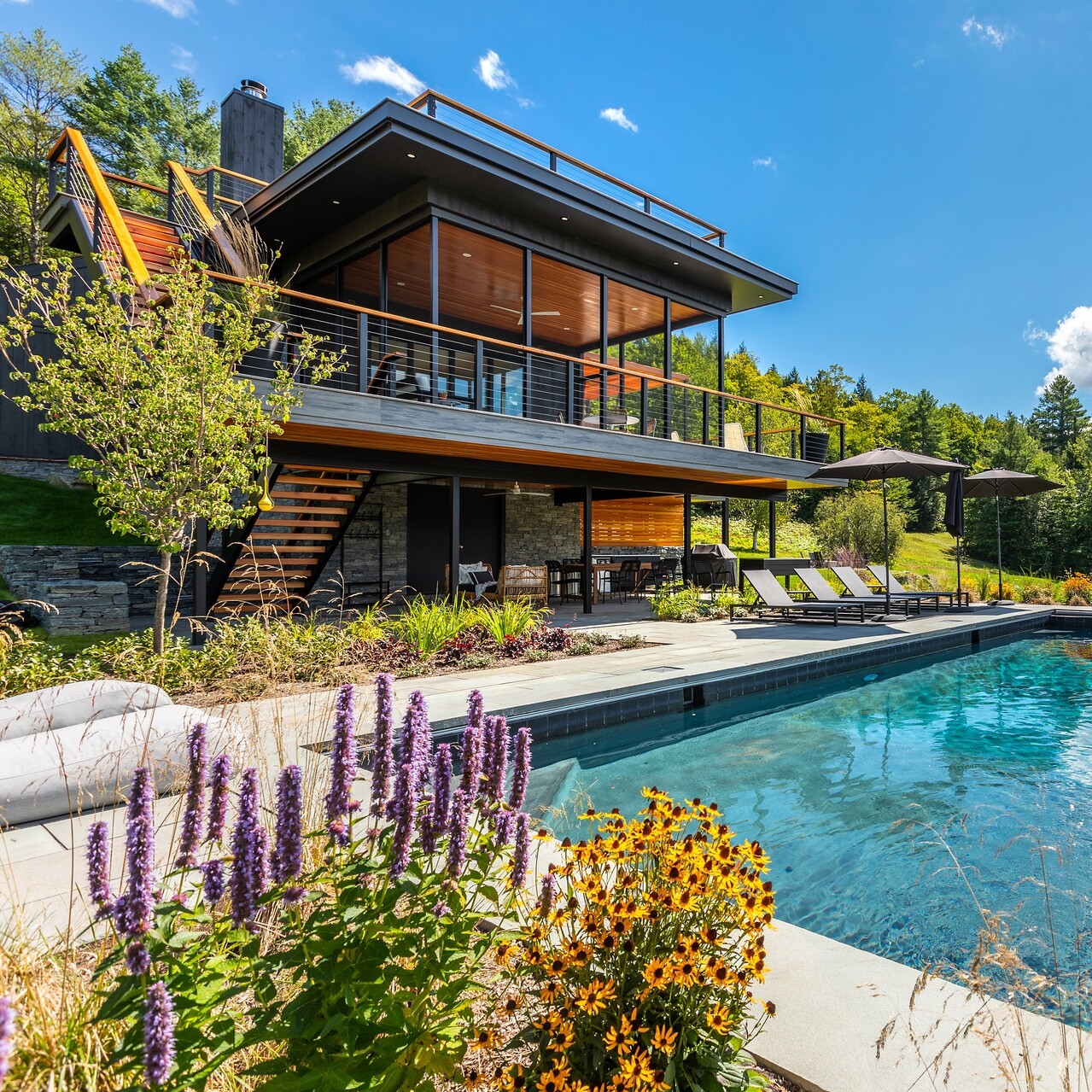 Modern house with a pool, vibrant flowers, and views of greenery under a clear blue sky.