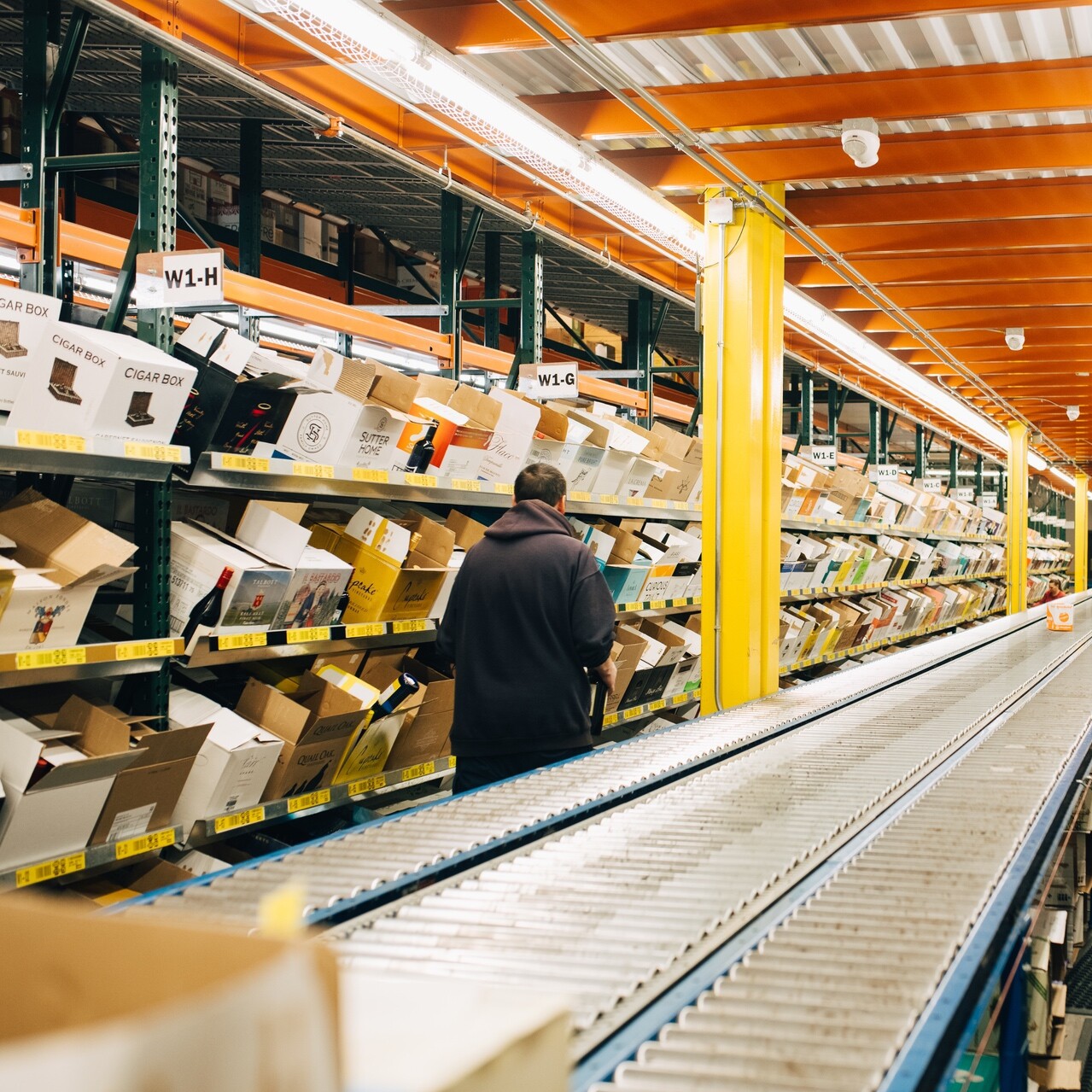 Warehouse aisle filled with boxes and a person inspecting shelving labels.