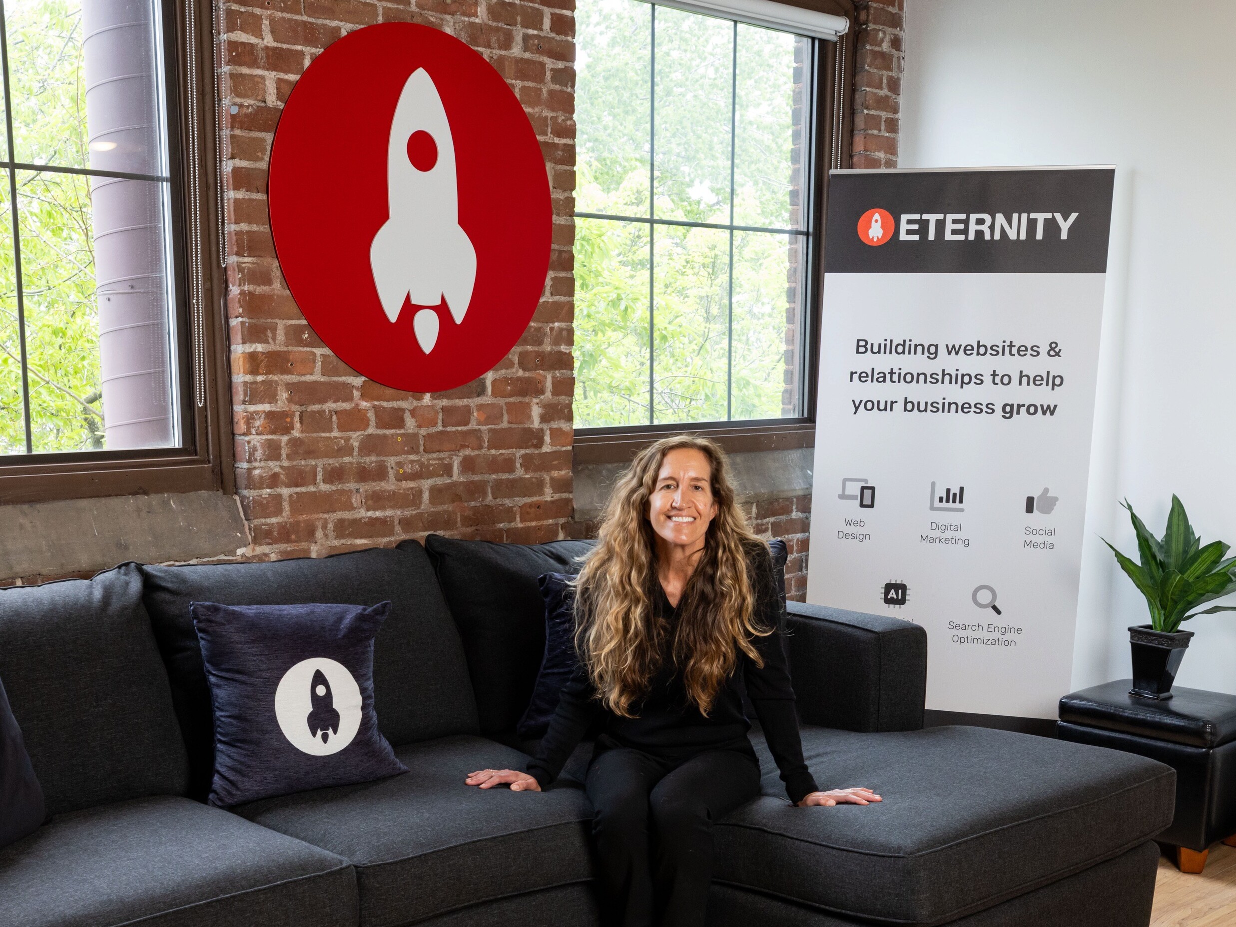 Woman sitting on a couch in an office with a rocket logo and a business banner behind her.