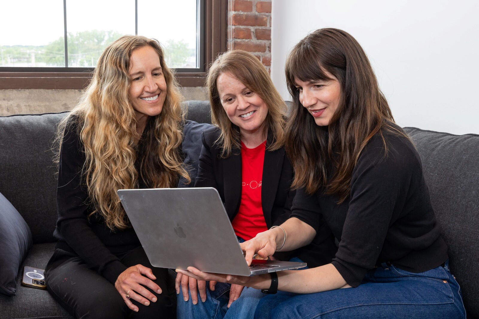 Three women are sitting on a couch, smiling and viewing a laptop together.