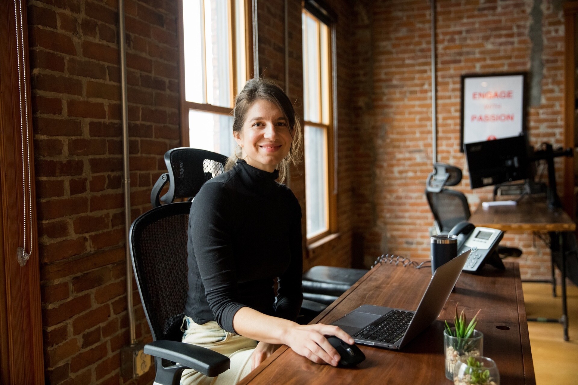 Smiling woman at a desk with a laptop, in a brick office setting.