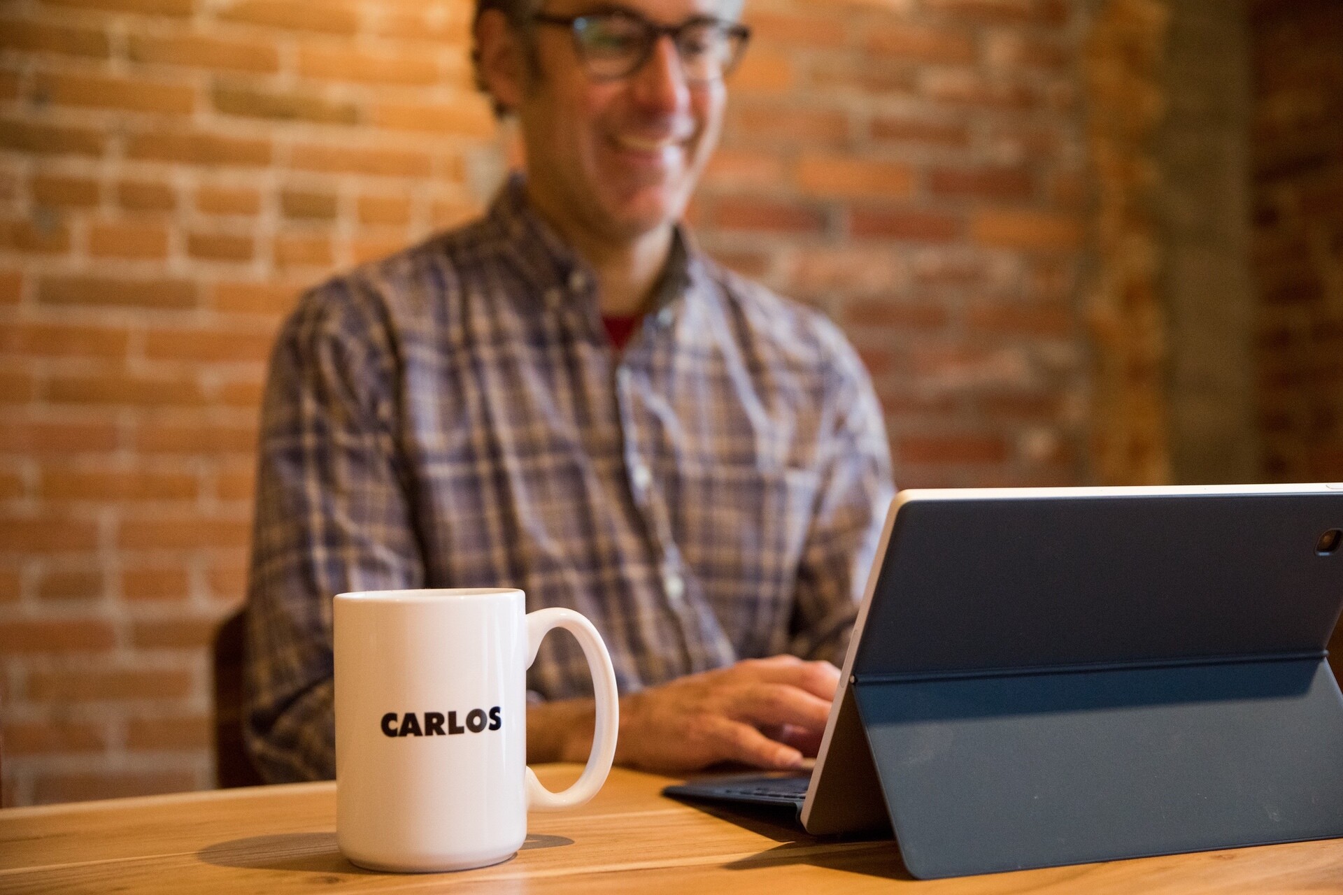 Man in plaid shirt smiles while working on a tablet; a coffee mug labeled "CARLOS" is visible.