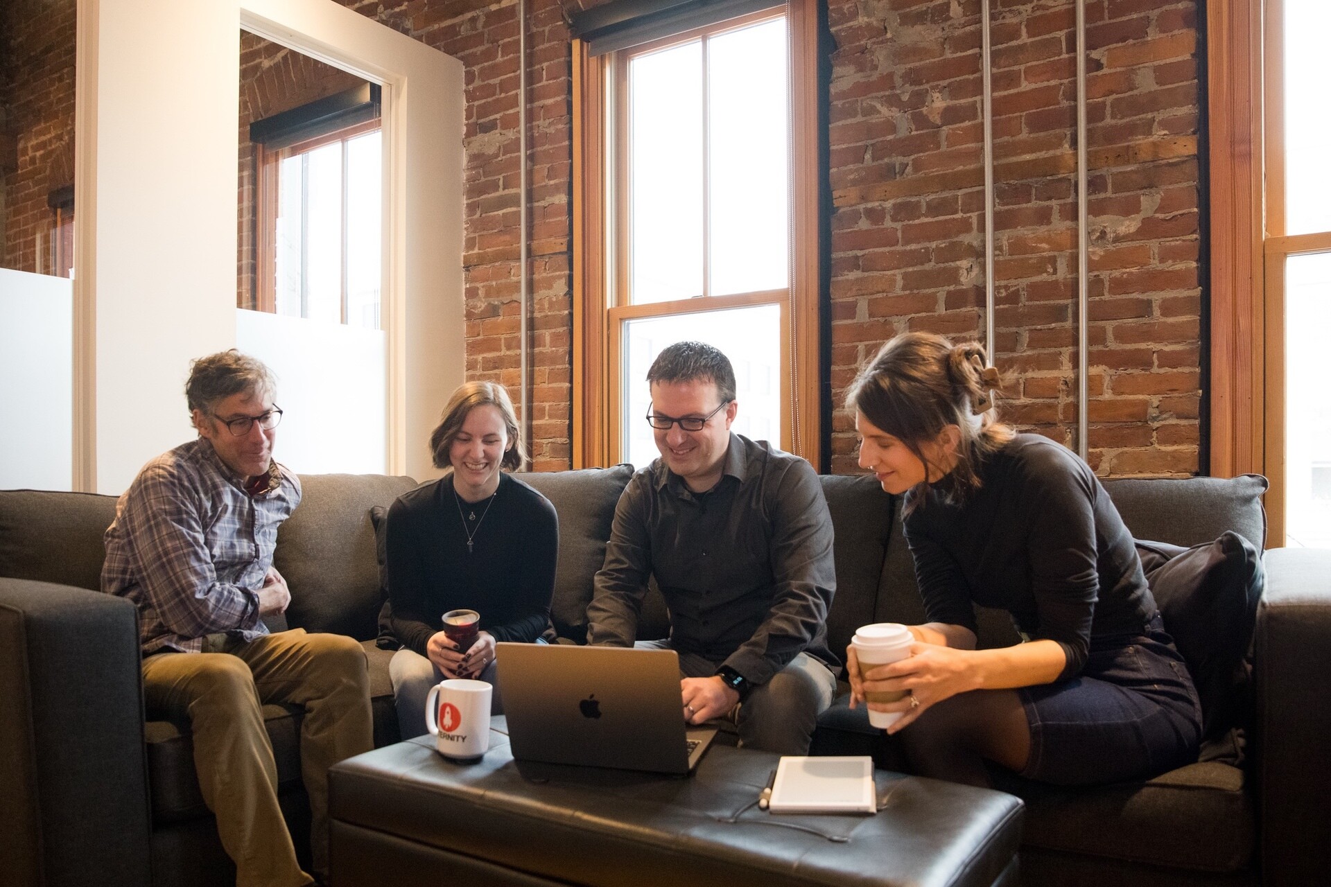 Four people sit on a couch, looking at a laptop and laughing, in a cozy brick-walled room.