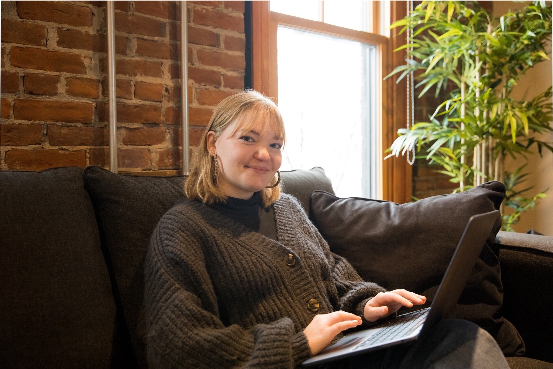 Young woman smiling while using a laptop on a couch, with a brick wall and plants in the background.