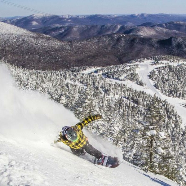 A snowboarder in a yellow plaid jacket carves down a snowy mountain slope.