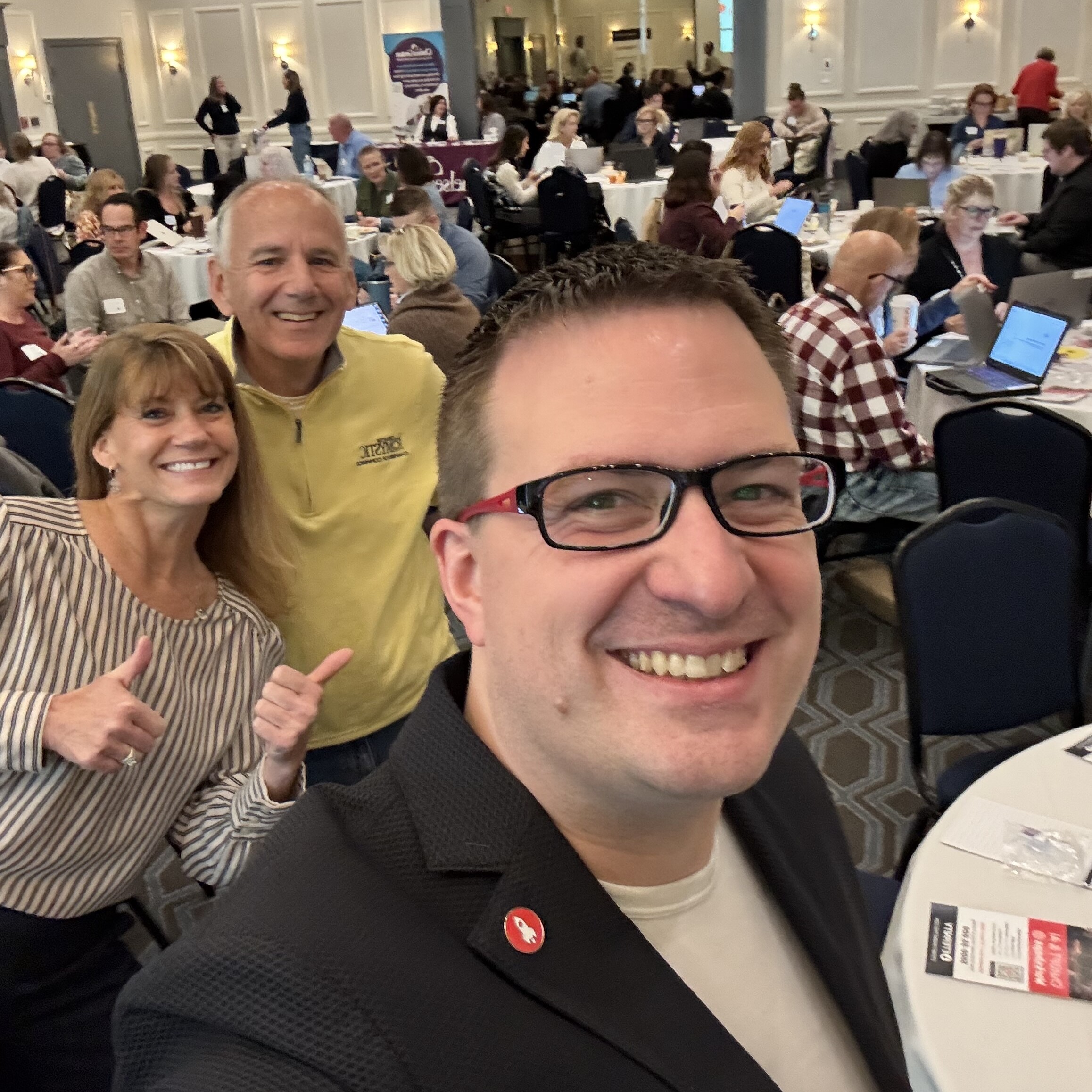 Three smiling people pose for a selfie in a conference setting with attendees in the background.