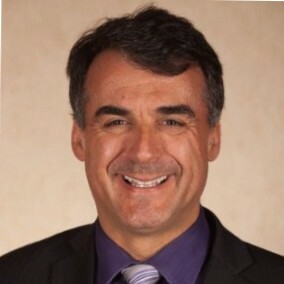 Smiling man in a suit with a purple shirt and tie against a light background.