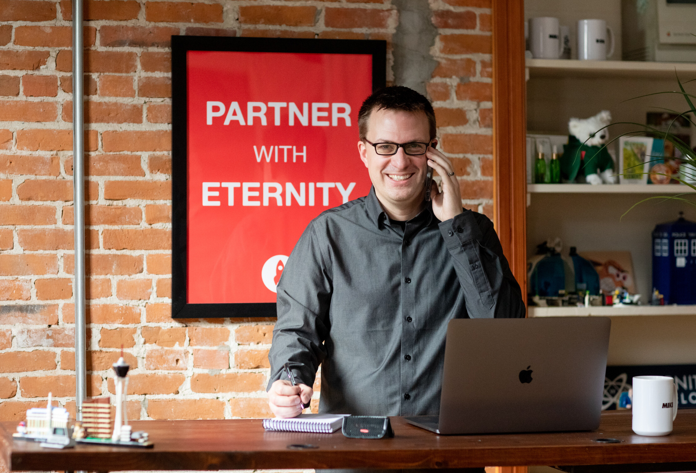 Man smiling on a phone call in an office with a "Partner with Eternity" sign.