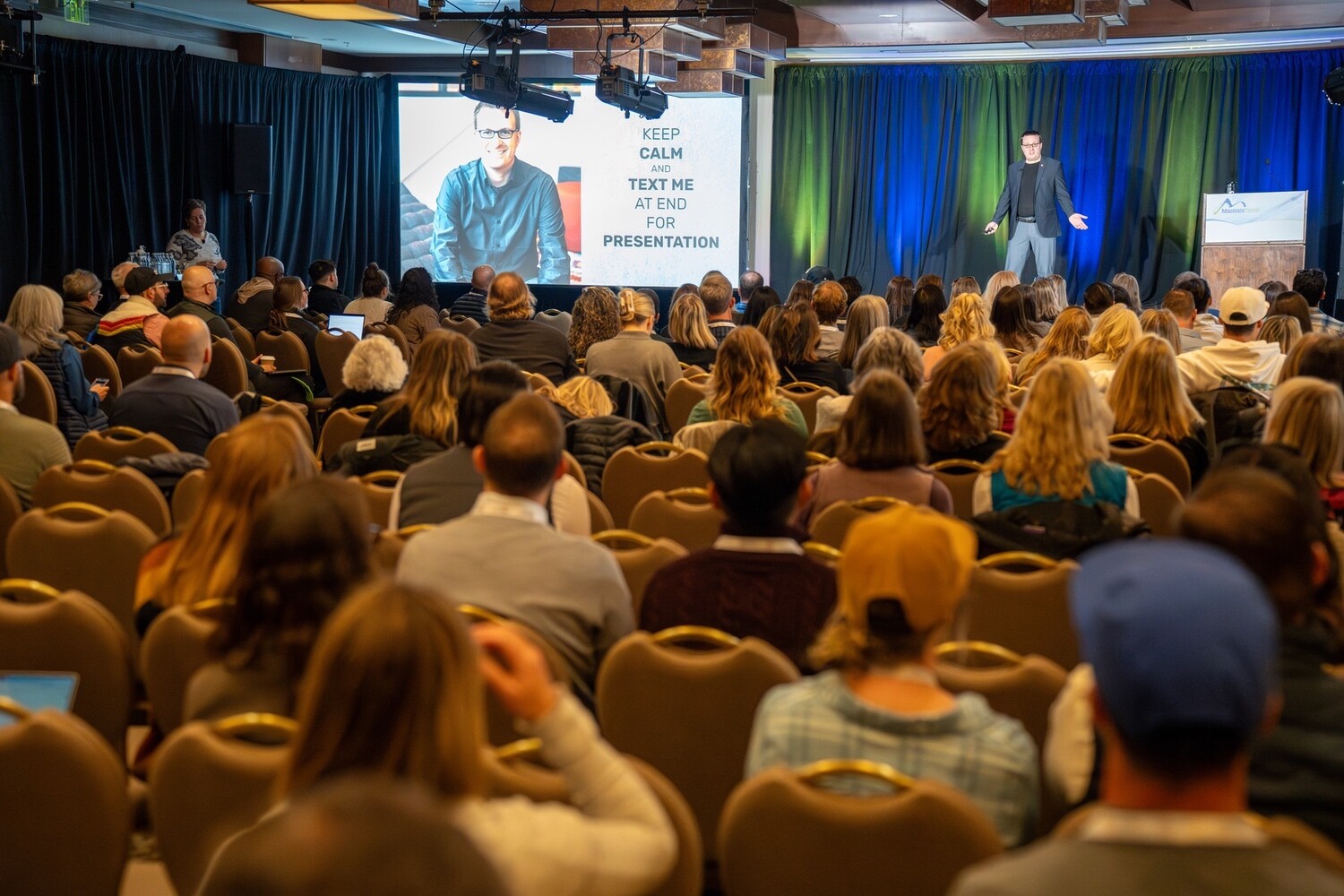 A speaker presents to an audience at a conference, with a slide displaying text behind them.