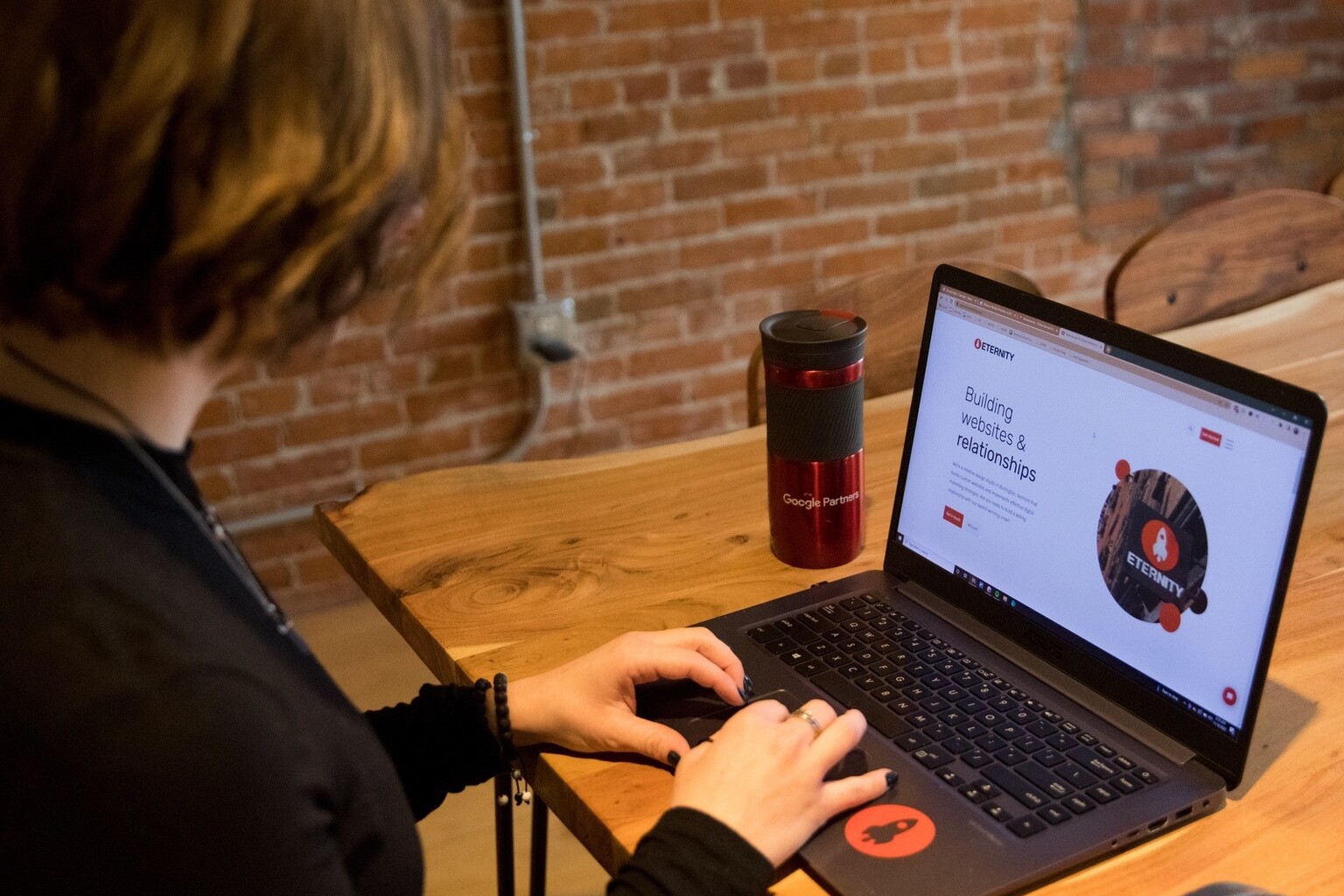 Person working on a laptop at a wooden table with a red travel mug nearby.
