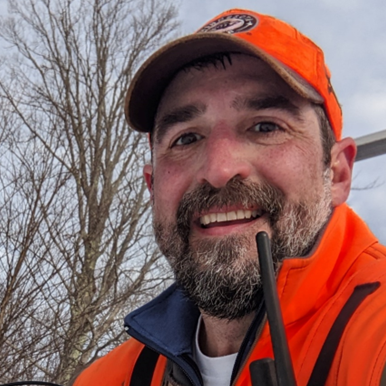Smiling man in an orange jacket outdoors, holding a radio with trees in the background.