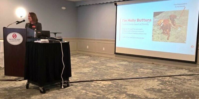 A woman speaks at a podium with a presentation screen behind her in a conference room.