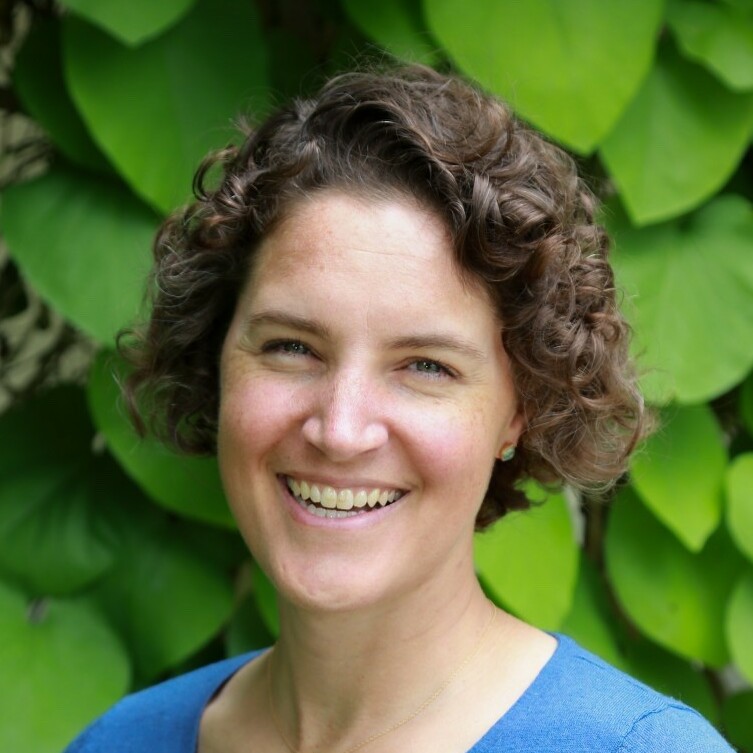 Smiling woman with curly hair in front of green leafy background.