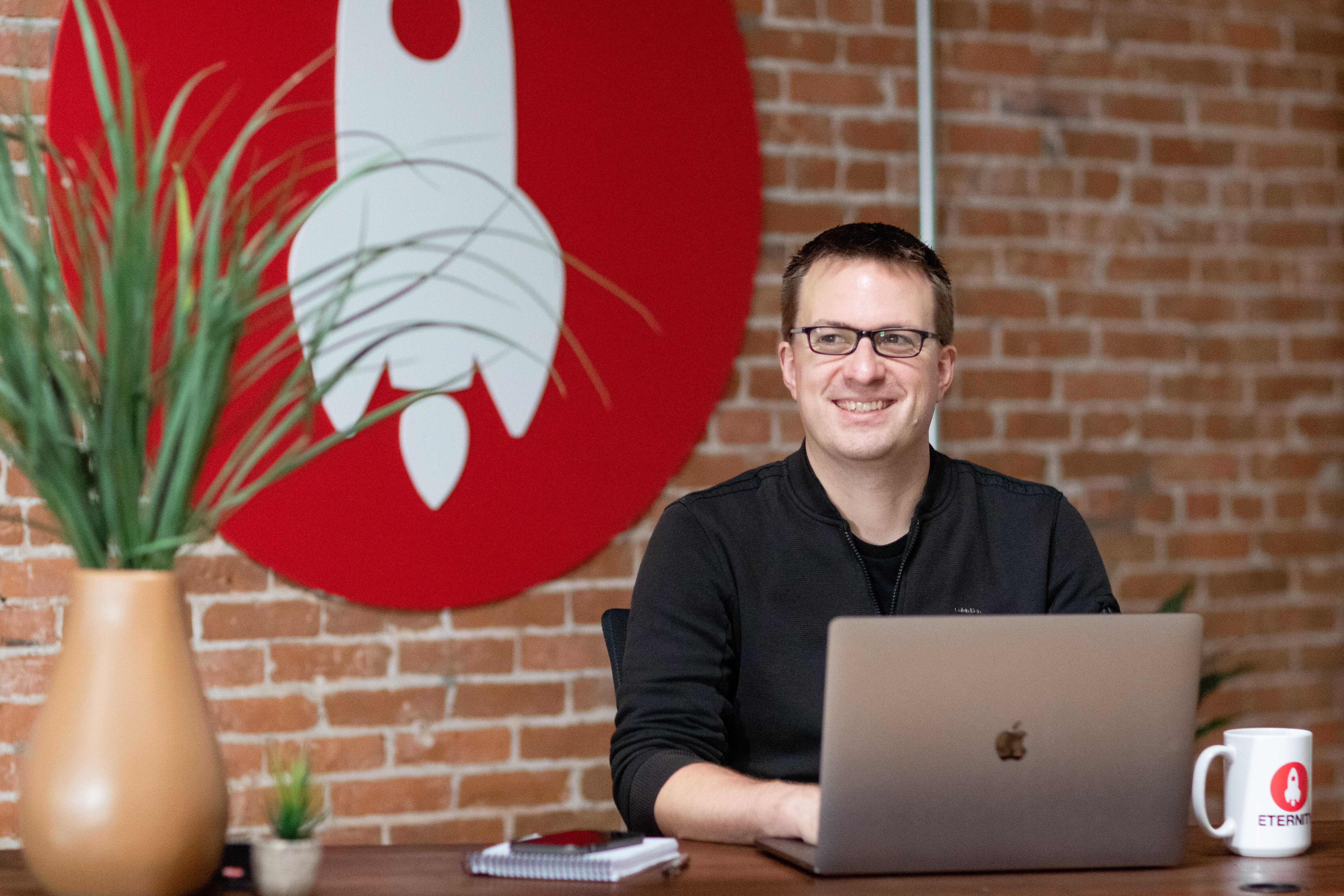 Person smiling at a desk with a laptop, plants, and a logo on the wall.