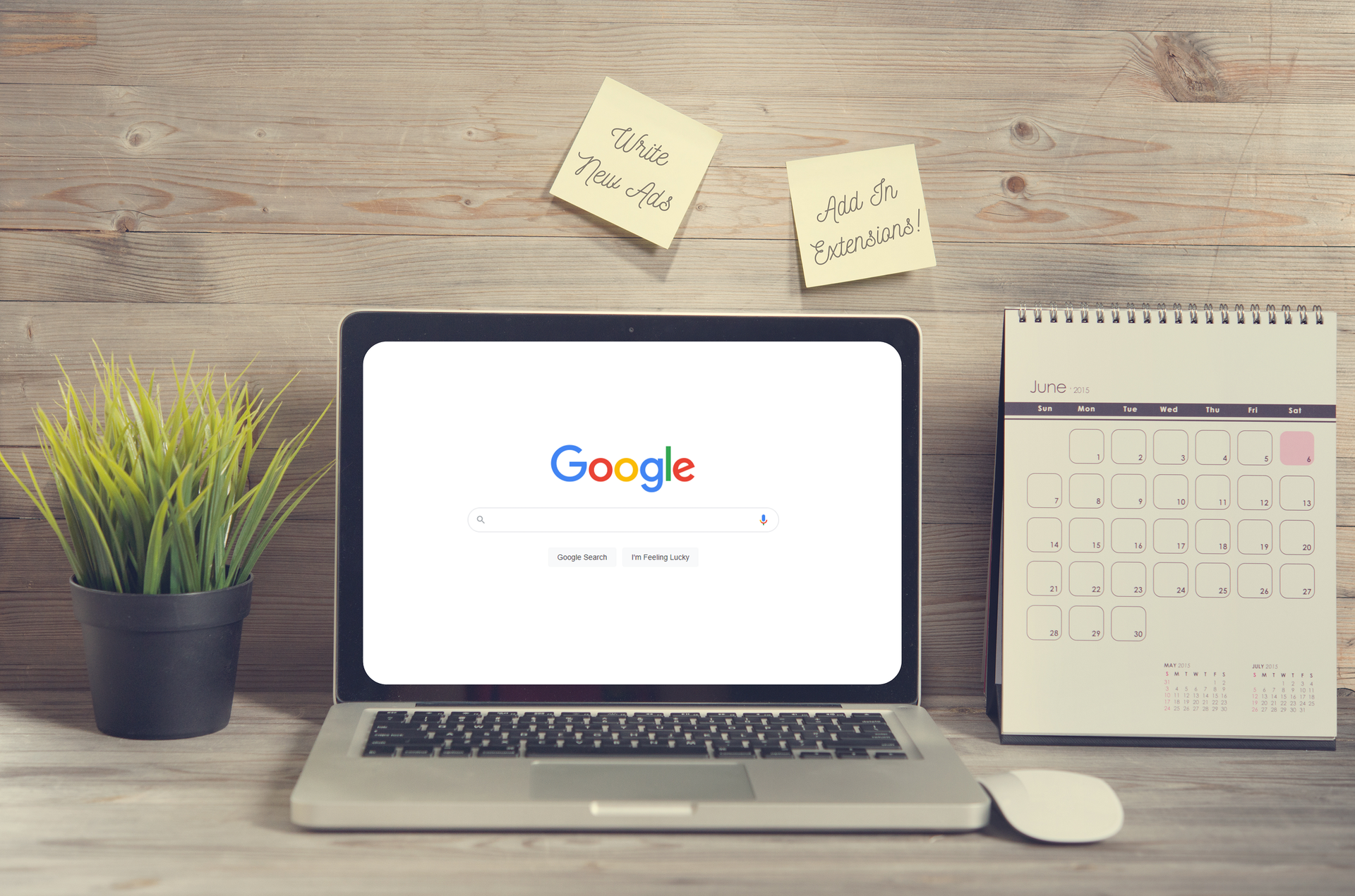 A laptop open to Google Search with a plant nearby and a calendar to the right of it. Sticky notes above referencing google ad extensions