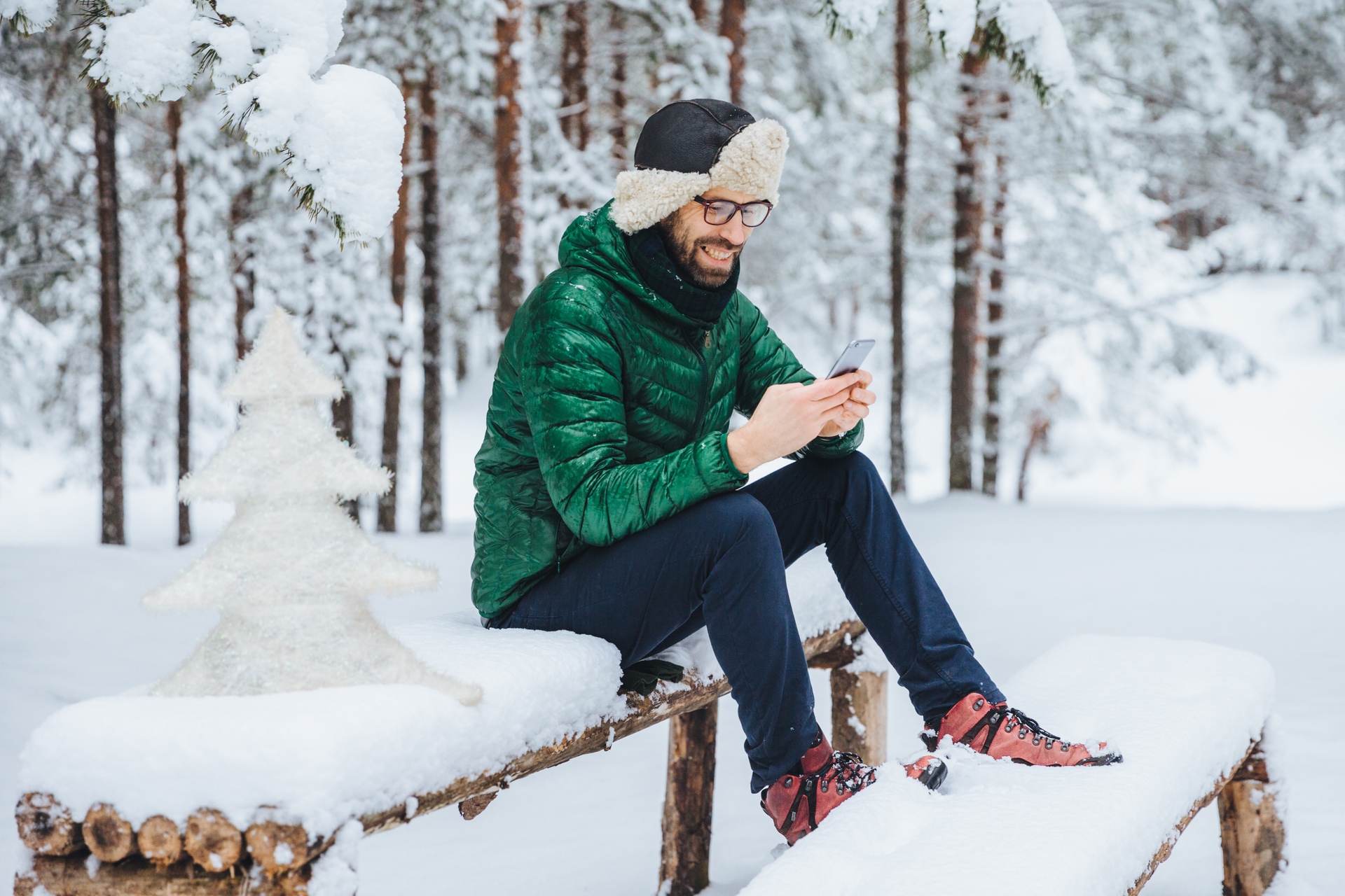 Happy man sitting on top of a picnic table covered in snow, next to an ice sculpture, wearing a hat with ear flaps and a green parka smiling at his phone in the middle of some snow-filled woods