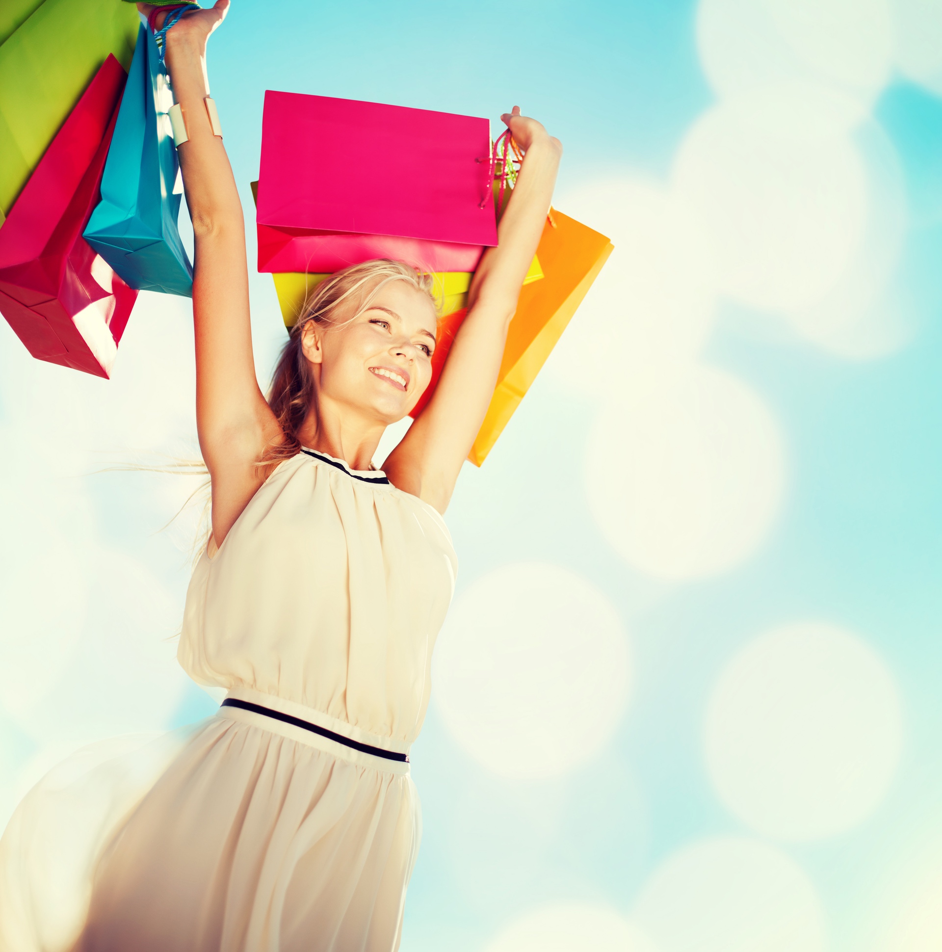 young blond girl smiling and holding multiple bags in different, cheerful colors above her head against a background of blue sky and soft clouds