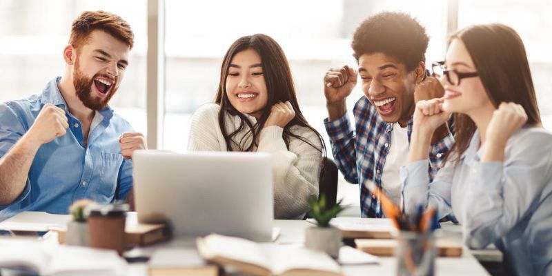 Multi-ethnic group of individuals in a friendly business meeting, smiling enthusiastically around a table with an open laptop and books.