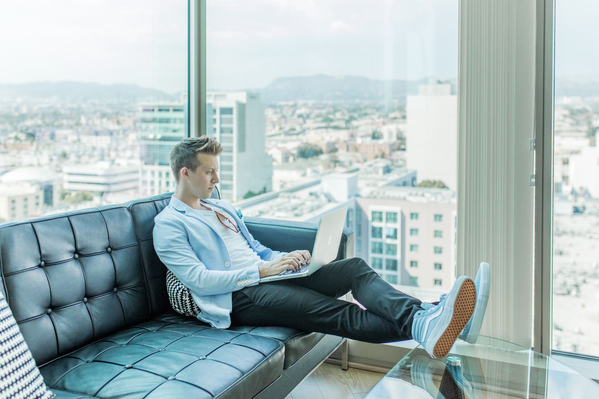 Guy on a leather couch with his feat on a table using a laptop.