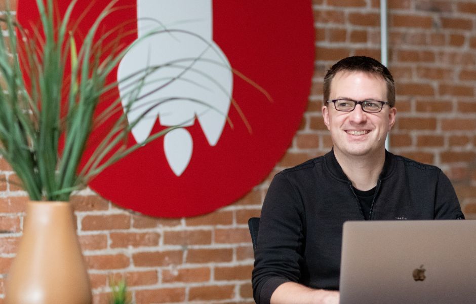 A person smiles while working on a laptop, with a rocket-themed wall behind them.